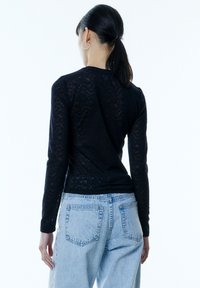 Schwarzes Langarmshirt mit einem Spitzenmuster, kombiniert mit hellblauen, hoch taillierten Jeans. Einfaches Design und enge Passform.