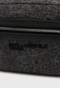 Saco de tecido cinza com uma superfície texturizada, apresentando um fecho preto e o logótipo em relevo "KARL LAGERFELD JEANS" em letras pretas.