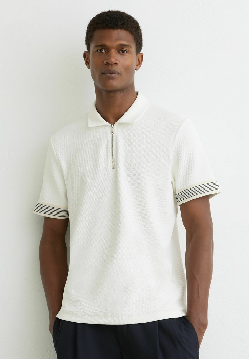 Ung mand iført en hvid kortærmet poloshirt med sorte stribede ærmekanter og sorte bukser, stående mod en hvid væg.