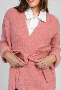 Cardigan en laine rose doux avec un design enroulé, doté d'une ceinture à nouer et de manches larges. La texture semble confortable, complétée par une chemise blanche à col en dessous.