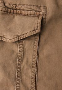 Beige látka s hladkým povrchem obsahuje náprsní kapsu se špičatým klopou, zesílené švy na bocích a jemné diagonální linie.