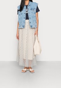 Spijkerhalster over een marineblauwe t-shirt, gecombineerd met een polka dot maxi rok en witte, geknoopte slippers. Houdt een crèmekleurige handtas vast.