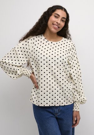 Blouse crème à pois noirs, décolleté rond, et longues manches bouffantes avec des poignets froncés. Associée à un jean bleu taille haute.