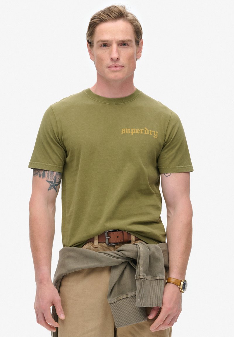 Olivengrøn bomulds-t-shirt med rund halsudskæring og korte ærmer, med et struktureret guldlogo. Brun bælte og khakifarvede bukser med en sweater bundet om livet.