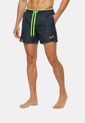 Uomo che indossa pantaloncini da bagno neri con coulisse verde neon e logo, in piedi a piedi nudi con una mano in tasca.