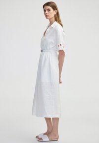 Witte linnen jurk met een kraag en korte mouwen, voorzien van uitsnijdingen. De jurk is enkel-lang en heeft een aansluitende taille, gedragen met witte slidesandalen.