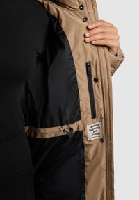 Tanová prošívaná bunda, zipová kapsa uvnitř, černá podšívka, nastavitelný šňůrkový pás, uvnitř přišitá látková etiketa s textem.