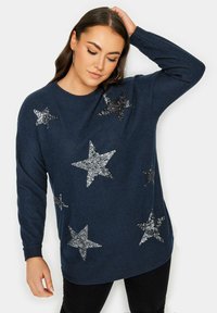 Marinblå stickad tröja med silverfärgade paljetter i stjärnmönster, rundad halsringning och långa ärmar. Materialet verkar mjukt och har en texturerad yta.