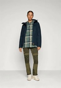 Mulher em pé, vestindo uma jaqueta azul-marinho sobre uma camisa xadrez verde e bege, calças verde-oliva com remendos reforçados e sapatos de caminhada bege-claro.