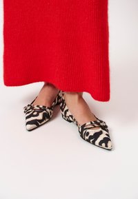 Puntige ballerina's in een zwart en beige dierenprint stof met kruisende bandjes en een decoratieve accent op de wreef. Rode gebreide rok erboven.