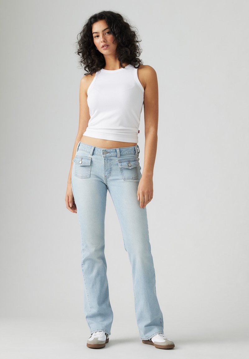 Jean flare en denim bleu clair avec poches avant et accessoires en argent, associé à un haut sans manches ajusté blanc. Texture lisse, style décontracté.