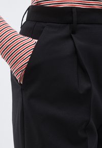 Χέρι στην τσέπη μαύρου ψηλόμεσου πλισέ παντελονιού, φοράει μακρυμάνικη μπλούζα με λεπτές κόκκινες, μαύρες και λευκές οριζόντιες ρίγες.