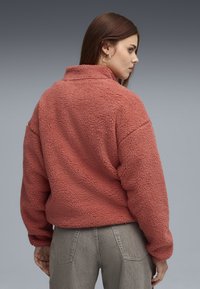 Pullover in pile di colore corallo caldo, con colletto alto, superficie texture e vestibilità ampia. Abbinato a pantaloni grigi chiari.