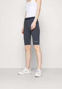 Šedé cyklistické šortky s bílým vyšitým logem na levé noze, v kombinaci s bílým tílkem a světlými sportovními botami.