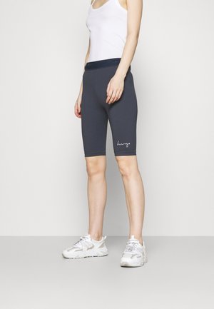 Grijze fietsshorts met een in het wit geborduurd logo op het linkerpand, gecombineerd met een witte tanktop en lichte sportschoenen.