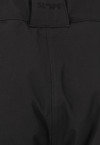 Pantalons en tissu noir avec coutures surpiquées, arborant un logo à la taille. Texture lisse, conçus pour la durabilité et une utilisation en extérieur.