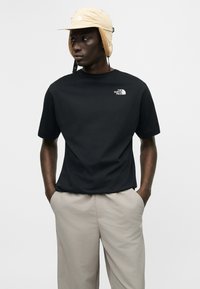 Άνδρας που φοράει μπεζ καπέλο φλοκάτης, μαύρο μπλουζάκι The North Face και ανοιχτό μπεζ παντελόνι, στέκεται με τα χέρια στις τσέπες κοιτάζοντας προς τα αριστερά.