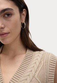 Beige stickad väst med V-ringning och flätad detalj. Har ribbade kanter och en mjuk textur. Modellen bär silverfärgade hoopsörhängen.