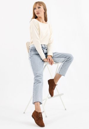Brown suede ankle boots with laces, cream long-sleeve top, and light blue jeans with frayed cuffs, seated on a white stool.