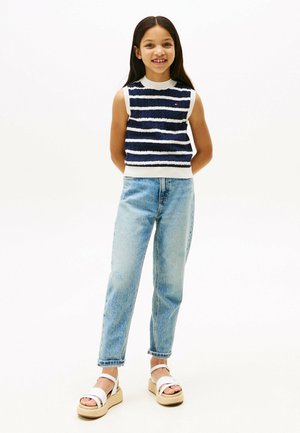 Top senza maniche a righe navy e bianche, jeans in denim blu e sandali con plateau bianchi caratterizzati da una suola texturizzata. La ragazza è in piedi con le mani dietro.