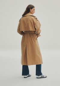 Cappotto trench marrone con vita a strisce, colletto oversize e spacco posteriore. Abbinato a jeans svasati blu e scarpe da ginnastica in bianco e nero.