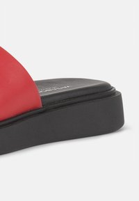 Røde syntetiske sandaler i slide-design med en bred stropp, glatt tekstur og en lav svart plattformsåle. Diskret logo-detalj på innersålen.