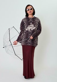 Übergroßer Leopardenmuster-Pullover in verschiedenen Lila- und Cremetönen, kombiniert mit einem bordeauxfarbenen, taillierten Rock und einem klaren Regenschirm.