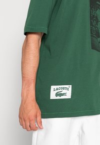 Camiseta de algodón verde con mangas cortas, que presenta una etiqueta rectangular blanca con el logo verde de "LACOSTE" y un gráfico de cocodrilo en el dobladillo.