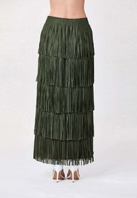 Jupe maxi verte à franges avec un design superposé, une finition texturée, une taille élastique et associée à des chaussures à talons hauts blanches.