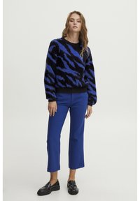 Črna in modra vzorčasta puhasta pulover z okroglo izrezi, v kombinaciji z enobarvnimi modrimi kratkimi hlačami in črnimi loaferji.