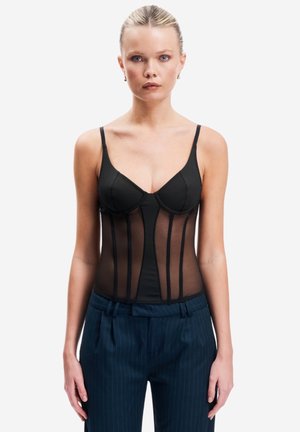 Femme aux cheveux blonds portant un corset noir transparent et un pantalon bleu marine à fines rayures, debout devant un fond blanc.