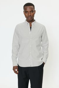 Mies, jolla on vaaleanharmaa raidallinen nappipaita, jossa ylin nappi auki, mustat housut, seisoo yhdellä kädellä taskussa.