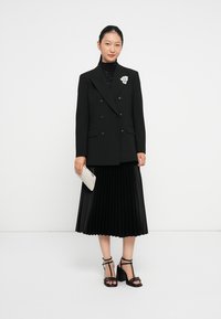 Blazer nero doppiopetto con rever a punte, abbinato a una gonna nera plissettata, mentre tiene una clutch bianca testurizzata. Sandali neri con tacco.