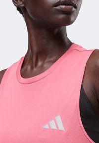 Gros plan d'une personne portant un haut de sport rose sans manches avec un logo gris Adidas sur la poitrine et un soutien-gorge de sport noir en dessous.