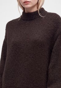 Femme portant un pull épais en tricot marron foncé avec un col haut côtelé, visible des épaules au menton sur un fond neutre.