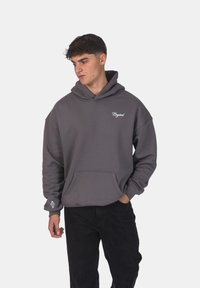 Felpa grigia in morbido materiale, con tasca frontale, cappuccio regolabile con cordino e logo ricamato bianco sul petto e sulla manica. Pantaloni neri.