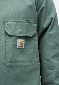 Πράσινο μπουφάν Carhartt σε κοντινό πλάνο που δείχνει μια κουμπωτή τσέπη στο στήθος με λευκή ετικέτα που φέρει το κίτρινο λογότυπο της μάρκας.