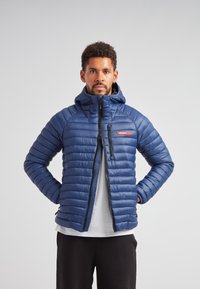 Blå puffjacka med huva, med kviltat mönster, en framzipper och en liten röd logotyp på övre vänstra bröstet.