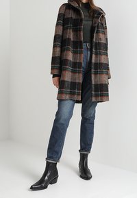 Rutet uldfrakke i brun, sort og grøn med løs pasform, knaplukning og stor krave; kombineret med blå jeans og sorte ankelstøvler.