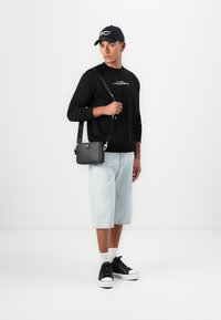 Sweatshirt preto com logo dourado, calções de ganga azul claro, mala preta de tiracolo e ténis pretos com solas brancas. Fundo branco.