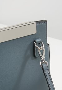 Blå läderhandväska med en slät textur, metallspänne och justerbar rem. Har en minimalistisk design och silverfärgade hårdvarudetaljer.