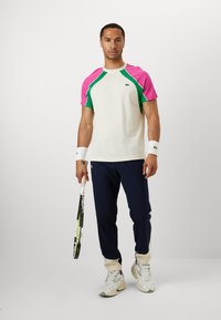 White sporty t-shirt with pink and green raglan sleeves. Paired with navy joggers and white sneakers. Holding a tennis racket.