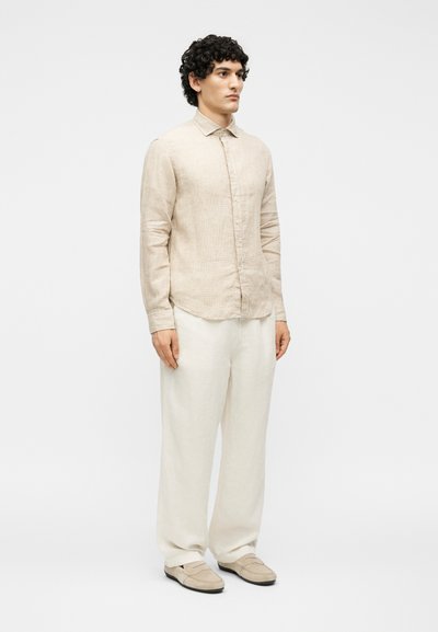 Άνδρας που φοράει μπεζ πουκάμισο με μακριά μανίκια και κουμπιά, λευκό φαρδύ παντελόνι και μπεζ μοκασίνια, μπροστά σε απλό λευκό φόντο.