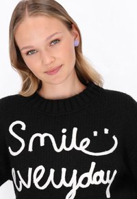 Junge Frau mit blonden Haaren trägt lila Ohrringe und einen schwarzen Pullover mit weißem Text, der "Lächle jeden Tag" und ein Smiley-Gesicht sagt.