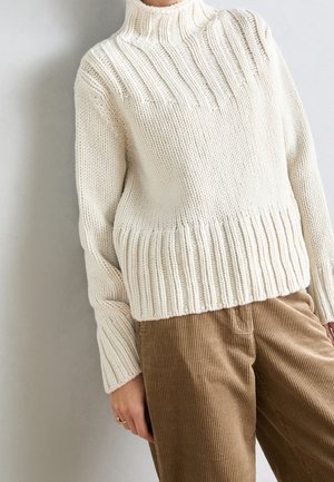 Pull en tricot crème avec col, poignets et ourlet côtelés, porté avec un pantalon en velours côtelé beige, main reposant le long du corps avec une bague en or.