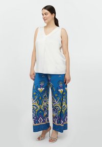 Biała bezrękawnik z dekoltem w kształcie litery V, w połączeniu z szerokimi niebieskimi spodniami o kolorowych wzorach kwiatowych i geometrycznych. Srebrne sandały na obcasie.