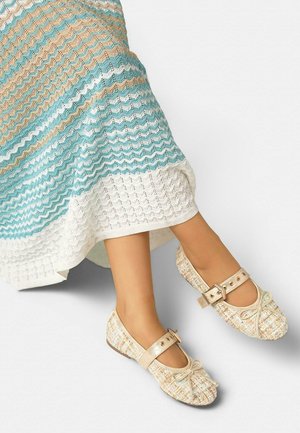 Picioare purtând o fustă tricotată multicoloră, asortată cu pantofi Mary Jane din tweed, cu curele cu catarame și fundițe decorative mici.