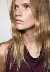 Gros plan d'une personne aux cheveux longs, ondulés, châtain clair, portant un haut sans manches bordeaux. Elle a les yeux bleus et un teint lisse.