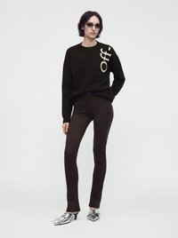 Femme aux cheveux courts et foncés portant un pull noir avec le texte blanc "Off-", pantalon sombre ajusté avec des bandes latérales, chaussures argentées à talons hauts pointus, et lunettes de soleil.