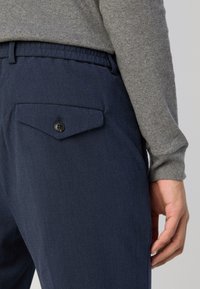 Granatowe spodnie o gładkiej fakturze, z tylną kieszenią zapinaną na czarny guzik. Na wierzchu widoczna jest szara koszula z długim rękawem.